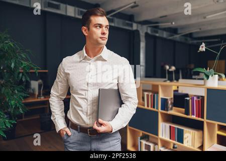 Portrait of attractive man manager standing with laptop on office background and looking side Stock Photo