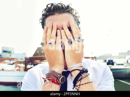 Hiding who he is...Close up portrait of a young man peeking through his fingers covering his face while standing on a wharf. Stock Photo