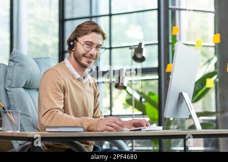 A young businessman man in a headset is concentrating on working from a home office at a computer. Smiling at the camera. Stock Photo