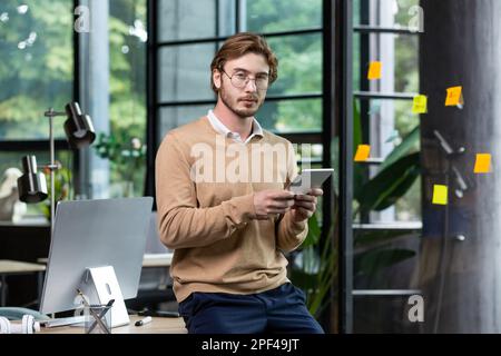 Portrait of a young male student, intern standing in the office, co-working at a table with a computer, using a tablet, studying online. Looks into the camera. Stock Photo