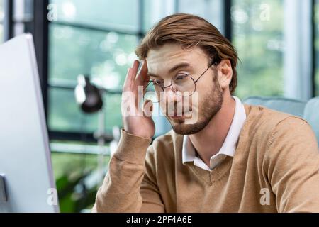 Close-up photo. Tired exhausted man, office worker, manager or freelancer sitting at his desk, tired of working on computer, overworked, having headache, need rest and break. Stock Photo