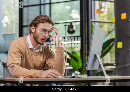 Tired exhausted man, office worker, manager or freelancer sitting at his desk, tired of working on computer, overworked, having headache, need rest and break. Stock Photo