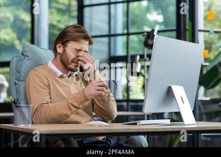 Tired male freelancer, programmer rubs his eyes, suffers from overfatigue, discomfort long sitting work in home office interior. Stock Photo