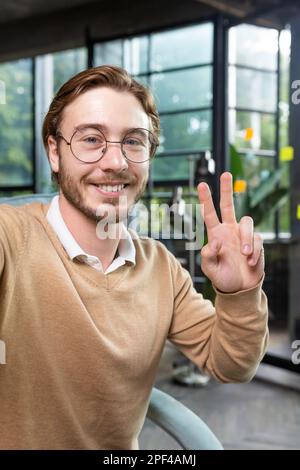 Close-up photo. Portrait of professional smart male programmer hacker taking selfie with hand, showing victory gesture with fingers. Smiles into the phone camera. Stock Photo