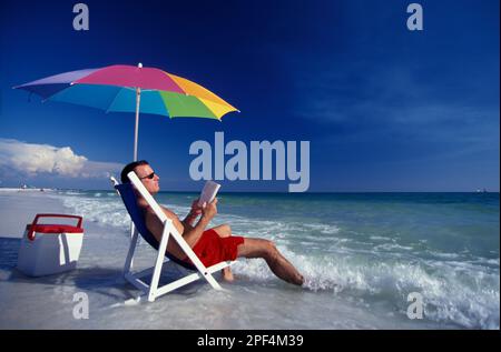 Caucasian men relaxing at the beach under a colorful and brother while reading a book Stock Photo