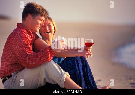 Young Caucasian couple enjoying a glass of wine well sitting barefoot on the beach Stock Photo