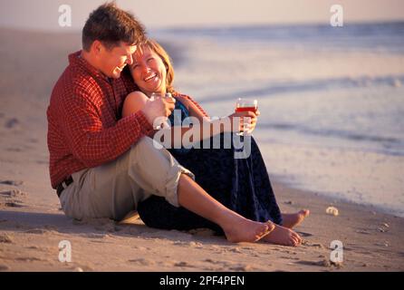 Young Caucasian couple enjoying a glass of wine well sitting barefoot on the beach Stock Photo