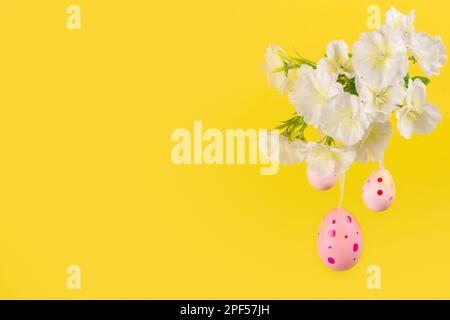 Captivating display of Easter cheer, depicting three pink eggs gently swaying from a leafy branch in bloom, set against a lively yellow hue background Stock Photo