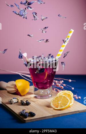 Summer purple drink/cocktail with petals and a yellow straw, lemon slices and ice, with colored background Stock Photo