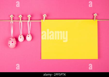 Blank yellow sheet of paper and three pink Easter eggs hang at rope on pink background. Easter greeting card concept with free space for writing Stock Photo