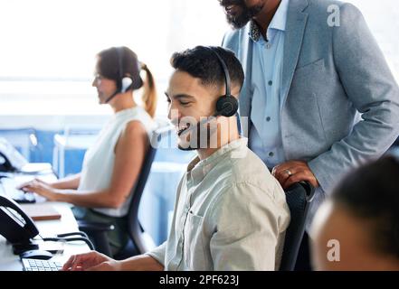 Youre doing a good job. a handsome young call centre agent sitting and using his computer while his manager watches. Stock Photo