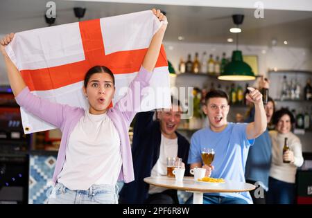 Company of young adult emotional fans supporting English team with state flag while resting in sports bar with beer Stock Photo