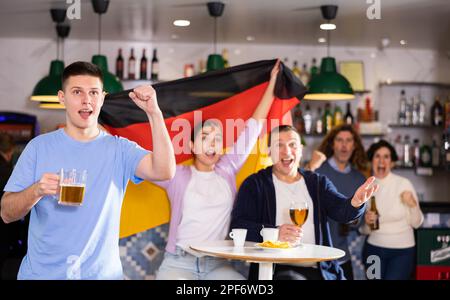 Company of emotional young adult fans supporting German team with state flag while drinking beer in sports bar Stock Photo