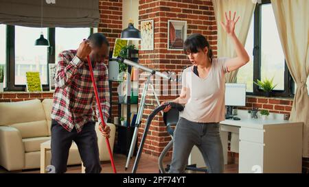 Happy life partners using tools to clean apartment, listening to music and dancing while they use mop and vacuum. Diverse couple cleaning living room floors, sweeping dust with cleaner. Stock Photo
