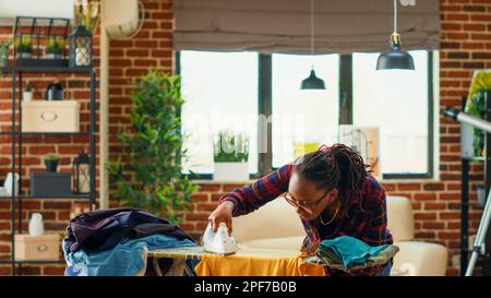 African american woman using steam iron and ironing board, smoothing out creases on clothes. Female person doing household chores for spring cleaning, using laundered clothing. Stock Photo