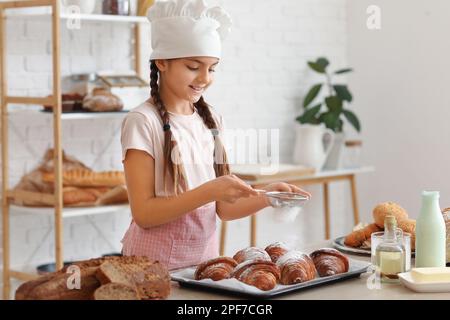 Little baker sprinkling sugar powder onto croissants in kitchen Stock Photo