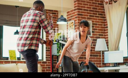 Cheerful modern people dancing and singing with cleaning tools, using mop and vacuum cleaner to wash floors. Young partners having fun with music while they clean apartment. Handheld shot. Stock Photo