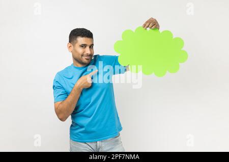 Cheerful unshaven man wearing blue T- shirt standing pointing at speech or think green bubble, empty communication cloud, looking at camera with smile. Indoor studio shot isolated on gray background. Stock Photo