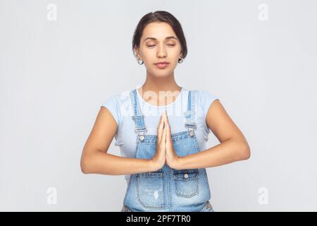 Portrait of calm relaxed young adult woman wearing denim overalls standing palm hands and doing yoga meditation with closed eyes. Indoor studio shot isolated on gray background. Stock Photo