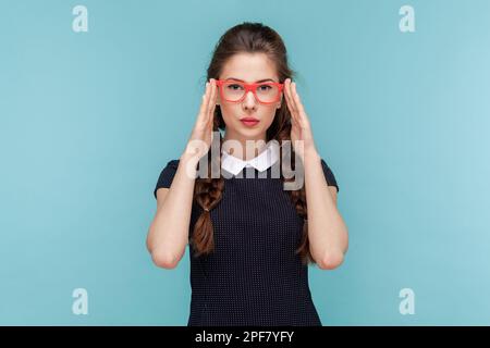 Portrait of serious attractive woman with braids stands keeps hands on frame of red glasses, looking at camera with attention, wearing black dress. woman Indoor studio shot isolated on blue background Stock Photo