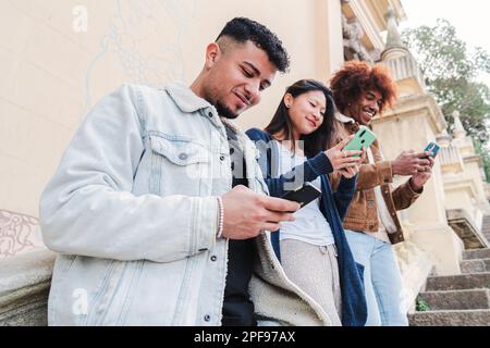 Multiracial group of three young friends enjoying and smiling using their mobile phone app. Diverse teenagers having fun sharing messages with each other on cellphone. Communication concept. High quality photo Stock Photo