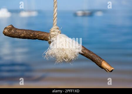 Close-up view of a stick tied to the end of a rope swing with blurred background of blue water and boats Stock Photo