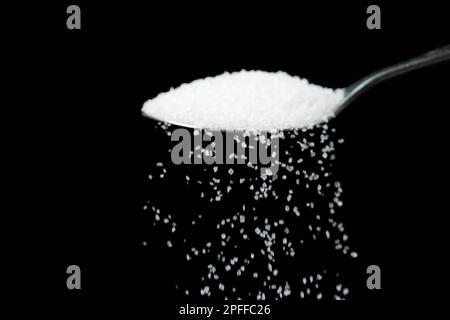 Macro photography of a teaspoon with white sugar on a black background, copy space. White sugar is poured from a teaspoon. Stock Photo