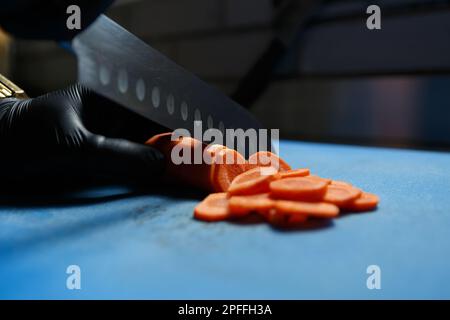 Cook wearing black gloves cuts fresh carrots with sharp santoku knife. Chef cutting vegetables on board in close up Stock Photo