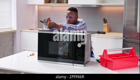 Happy Male Serviceman Using Screwdriver To Repair Microwave In Kitchen At Home Stock Photo