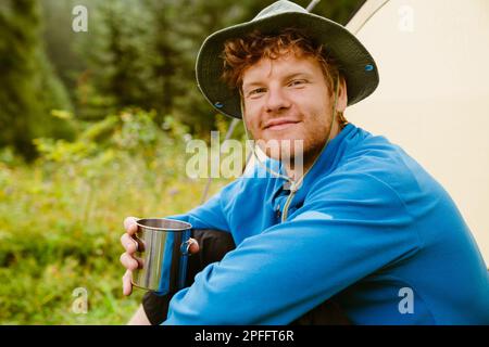 Young white tourist man drinking tea from metal cup while sitting at tent in forest Stock Photo