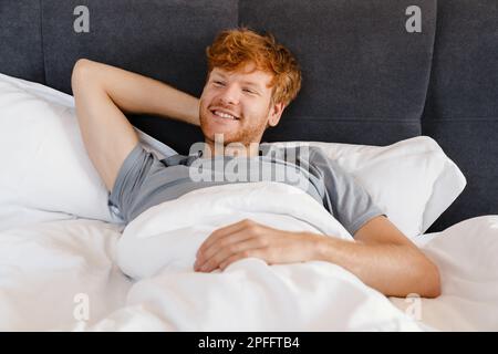 Young handsome smiling redhead man in gray t-shirt with hand behind his head looking aside lying on bed at home Stock Photo