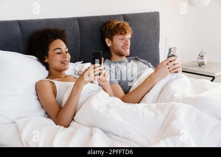 Young beautiful smiling interracial couple holding and using their phones, while lying in bed together. Handsome redhead guy and beautiful african cur Stock Photo