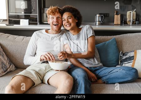 Young beautiful happy interracial couple watching cinema and eating popcorn, while sitting on couch in cozy sunny living room at home Stock Photo