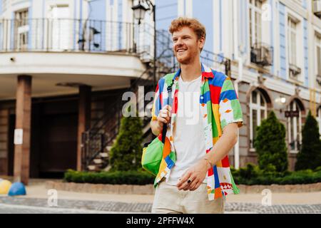 Young handsome redhead smiling man with stubble in colorful shirt walking street, near modern building Stock Photo