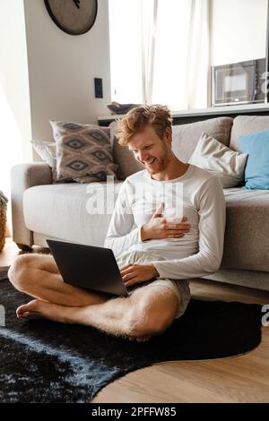 Young redhead man smiling and using laptop while sitting on floor at home Stock Photo