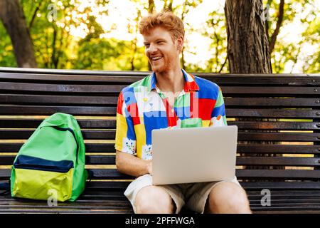 Young handsome smiling redhead man with laptop looking aside, while sitting with backpack on bench in park Stock Photo