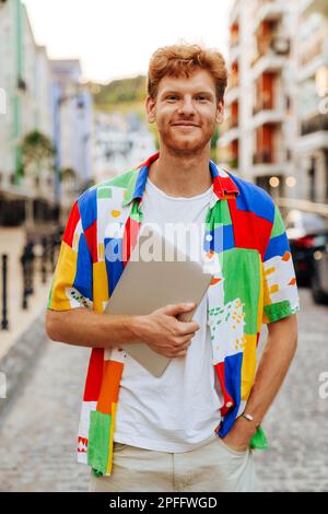 Portrait of young handsome smiling redhead man holding laptop and looking at camera, while standing on the street Stock Photo