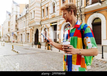 Portrait of young handsome calm redhead man with coffee holding and using his phone, while standing on the street Stock Photo