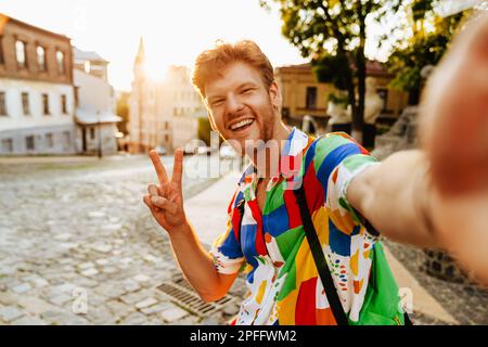 Selfie of young handsome smiling redhead man showing victory gesture with fingers, whuile standing on the street Stock Photo