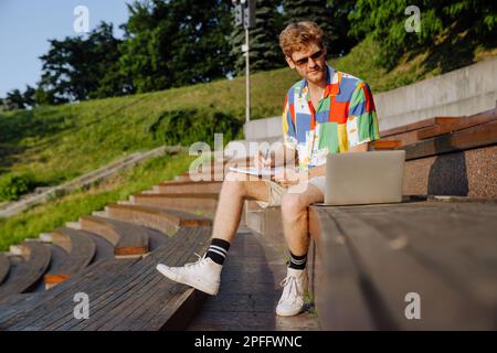 Young handsome stylish focused man in sun glasses doing homework with laptop, while sitting on tribune outdoors Stock Photo