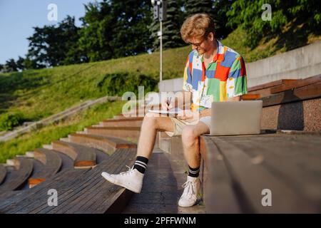 Young handsome stylish focused man in sun glasses taking notes , while sitting with laptop on tribune outdoors Stock Photo