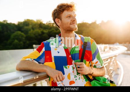 Portrait of young handsome smiling man in colorful shirt with backpack looking aside, while leaning on railing in the sun Stock Photo