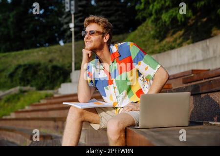 Young handsome stylish smiling man in sun glasses with laptop, textbook and pencil propping his head and looking aside, while sitting on tribune in su Stock Photo