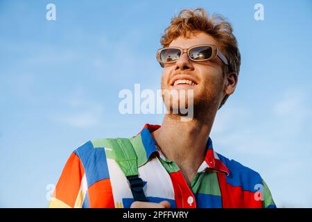 Portrait of young handsome stylish smiling man in colorful shirt and sun glasses looking aside Stock Photo