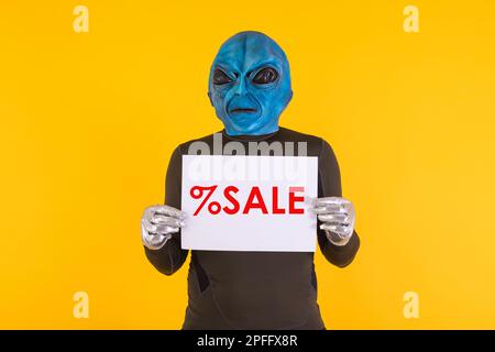 Man with an alien mask with a blue head, holding a white sign with red letters that reads: '% Sale'. Concept of bizarre, extraterrestrial, funny, info Stock Photo