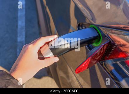 Hand plugging in the electric vehicle using the connector at the charging post, close-up shot. Stock Photo