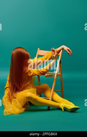 creative portrait of unknown redhead girl with long straight silky hair posing on armchair sitting in strange poses over cyan color background