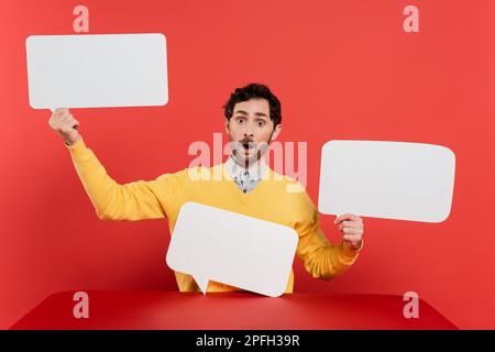 shocked man in yellow long sleeve jumper holding blank speech bubbles on red coral background,stock image Stock Photo