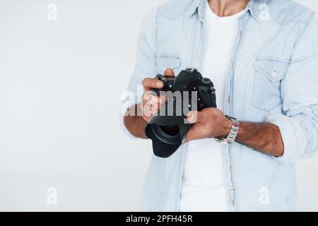 Holds camera in hands. Close up particle view of young man that standing indoors against white background Stock Photo