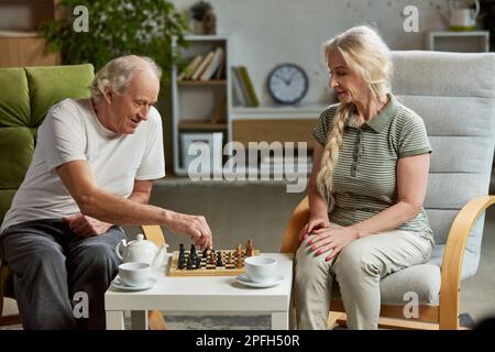 Happy elderly couple, retired man and woman spending time at home, playing chess together, drinking tea. Leisure time and hobby Stock Photo